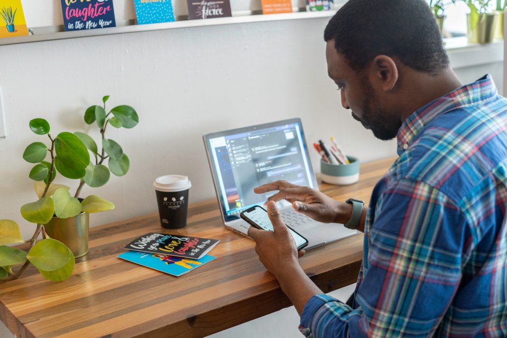 Un homme assis devant son bureau tient son smartphone en main tandis que son ordinateur portable est allumé