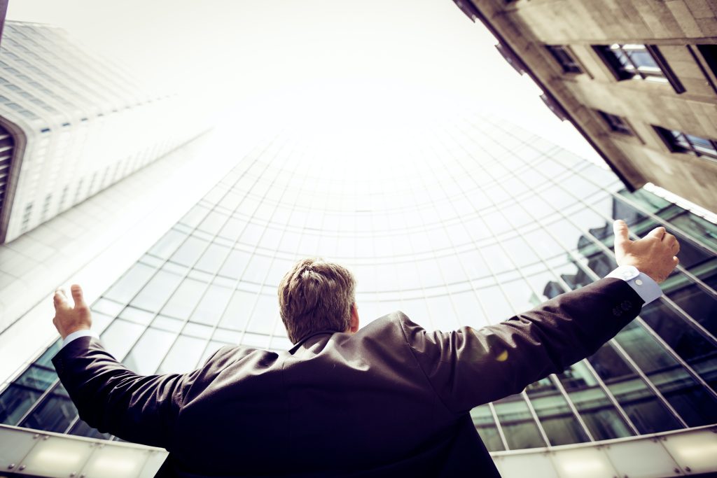 Un entrepreneur de dos est en train de lever les bras au ciel. La photo est en contre-plongée et il se trouve entre plusieurs grands immeubles d'affaires.