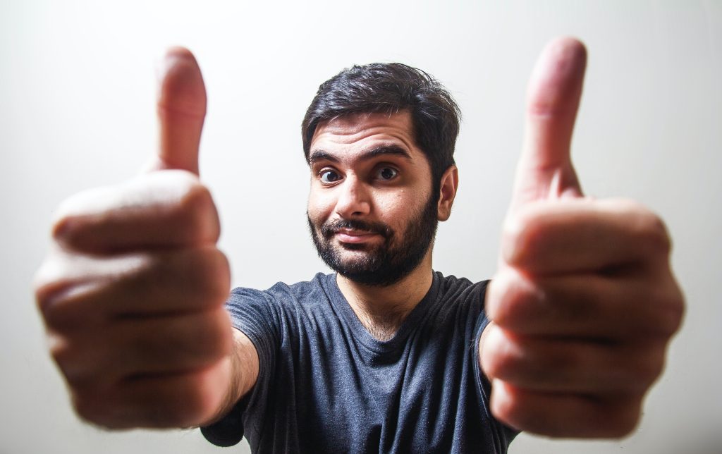 Un homme, de face et en gros plan, tient ses deux opuces levés près de l'objectif de l'appareil qui l'a photographié.