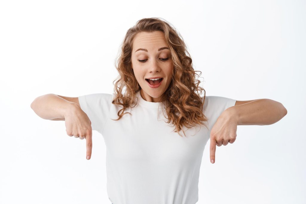 Une jeune femme blonde à l'air enjoué et vêtue d'un t-shirt blanc regarde vers le bas et montre du doigt vers le bas également