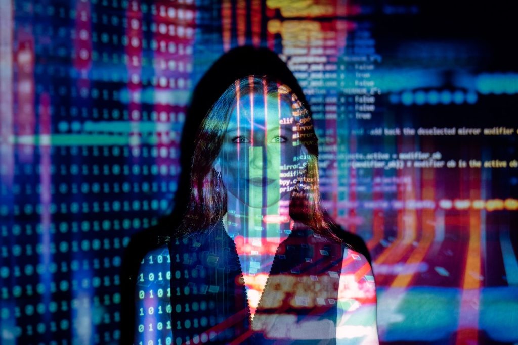 Une jeune femme se trouve dos à un mur sur lequel est projeté du code informatique de différentes couleurs.