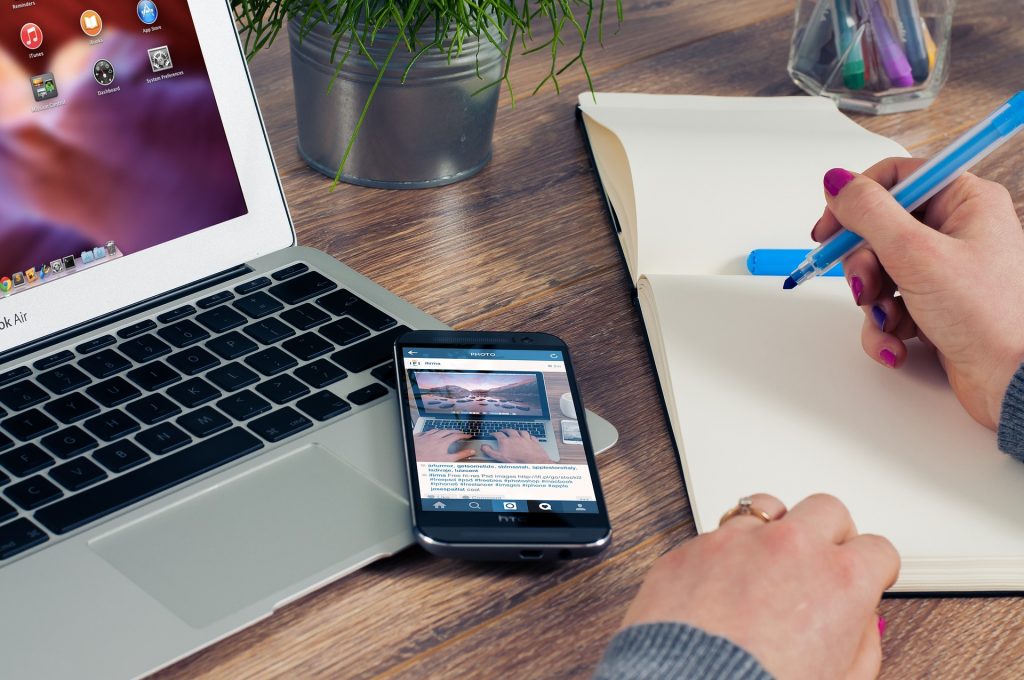 Une personne dont on ne voit que les mains écrit au feutre bleu dans un carnet posé à côté de son smartphone et de son ordinateur portable sur un bureau